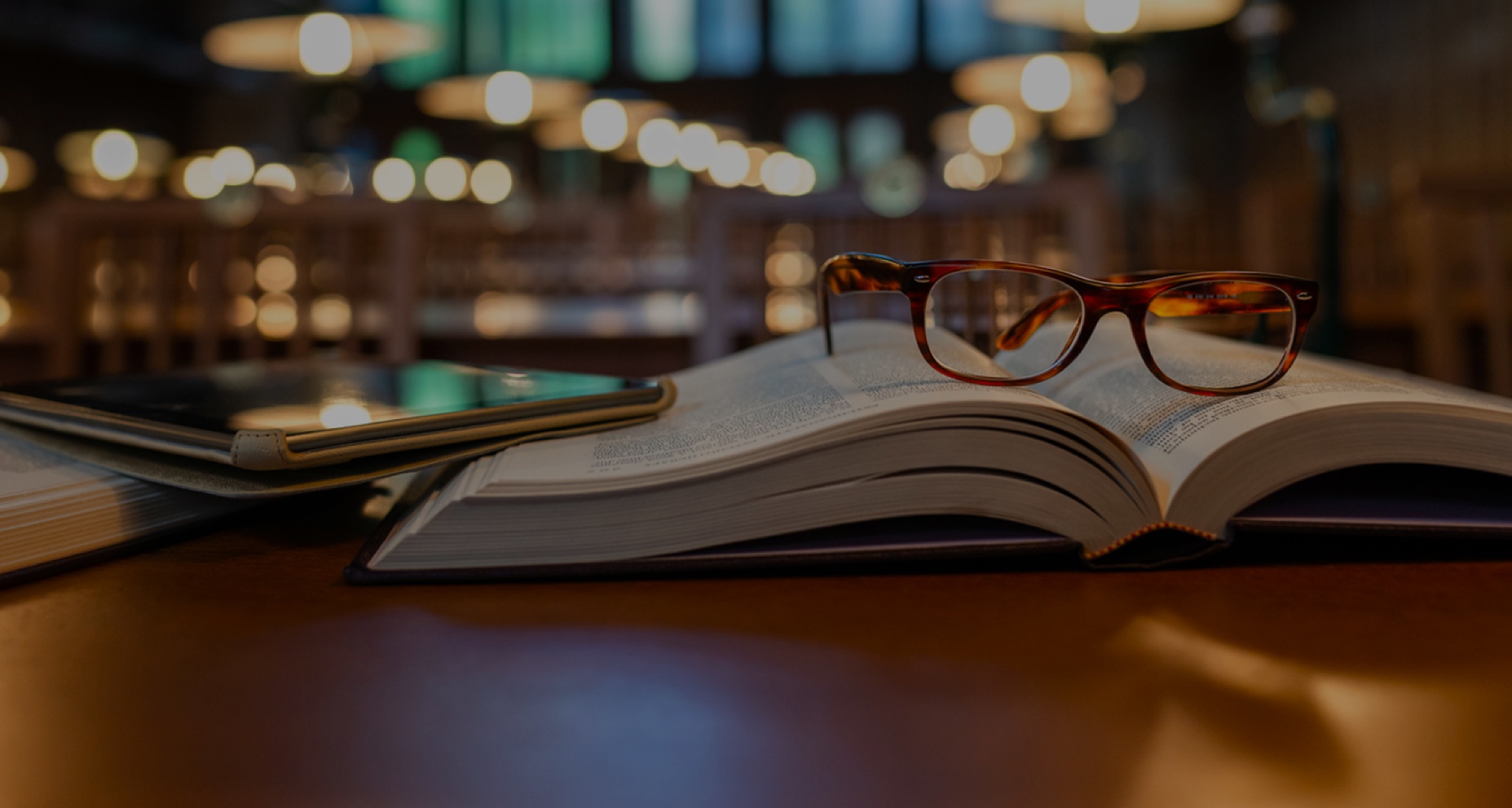A pair of glasses on top of a book and e-tablet on a desk in a library.