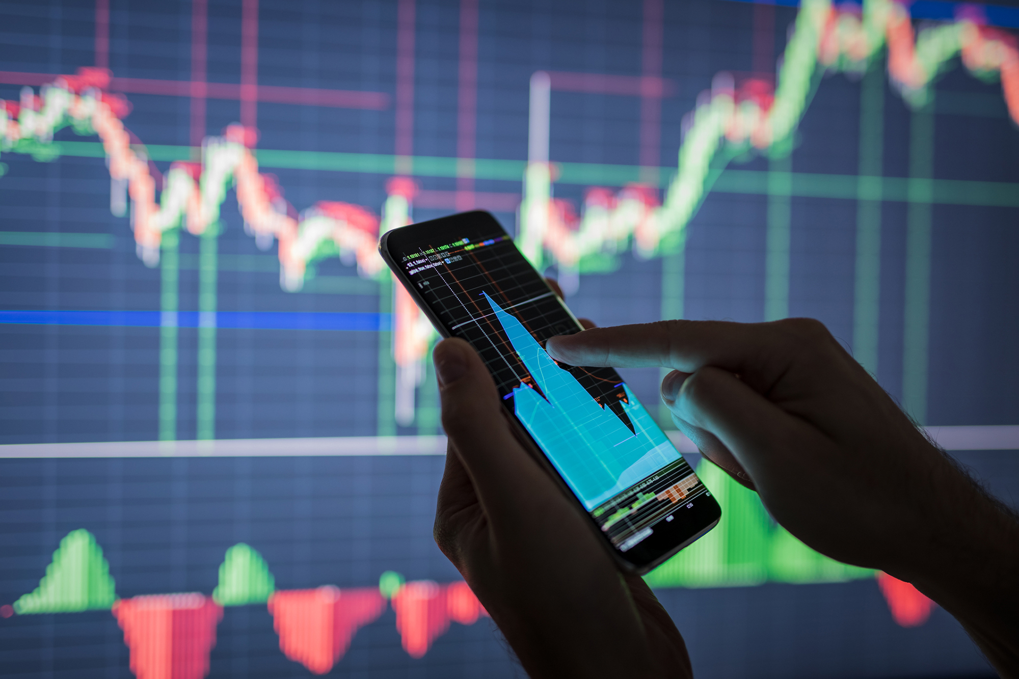 Business person checking market data on a mobile phone with data displayed on a screen in the background